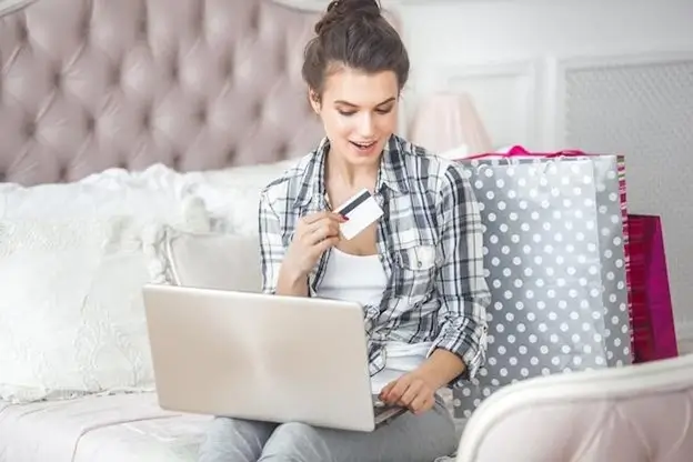 A woman sits on a bed, using a laptop while holding a credit card, engaged in online shopping or banking.