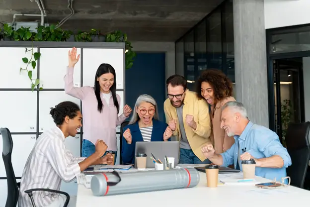 A diverse group of office workers raises their hands in celebration, showcasing teamwork and enthusiasm in a professional setting.