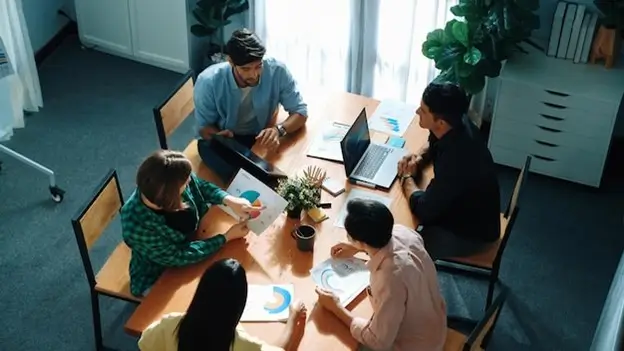 A diverse group of individuals engaged in discussion while using laptops at a round table in a collaborative setting.