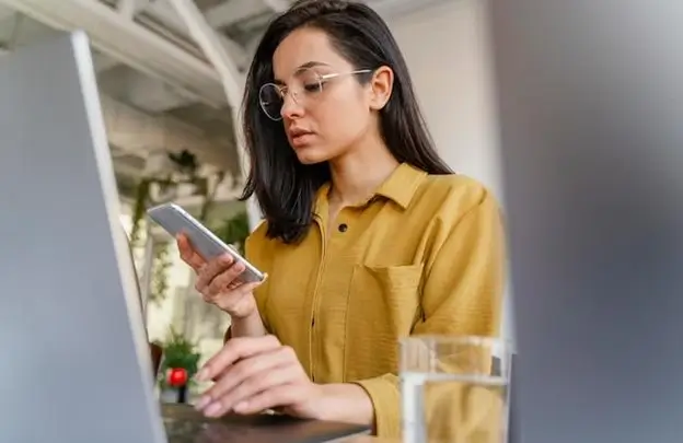 A woman wearing glasses is seated at a desk, focused on her phone while engaged in her work.