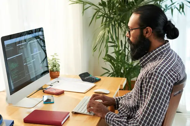 A bearded man wearing glasses sits at a desk, focused on his computer screen in a well-lit workspace.