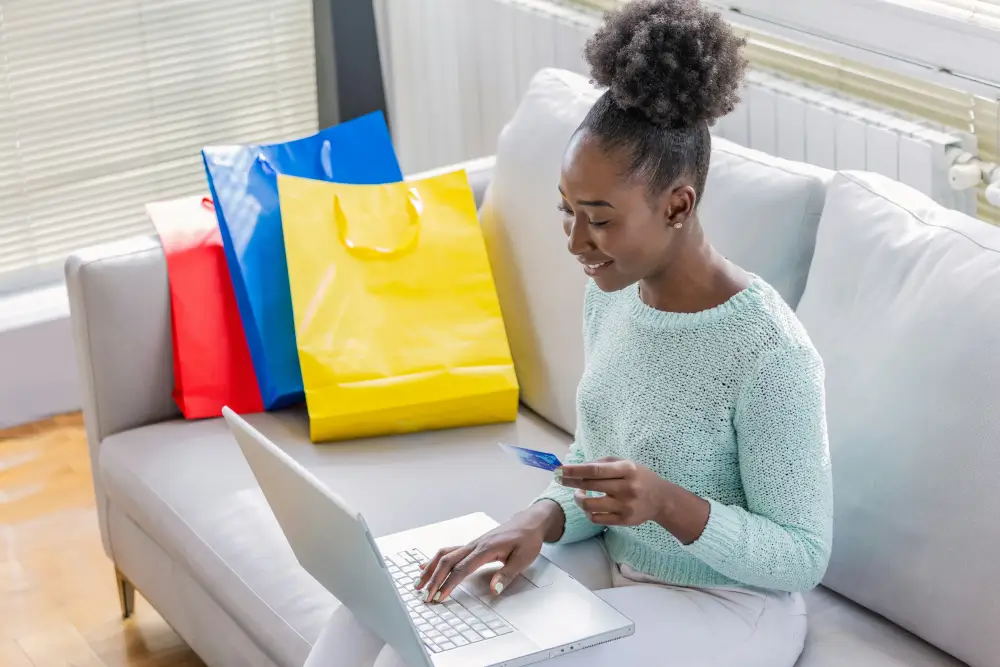 A woman types on a laptop, with shopping bags nearby, indicating she is shopping online.