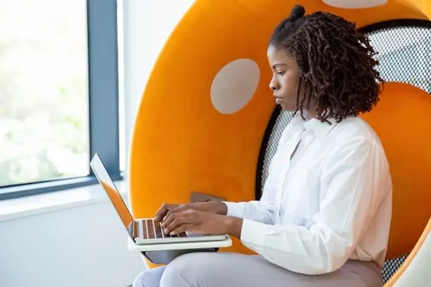 A woman seated in an orange chair, focused on her laptop, creating a productive and comfortable workspace.