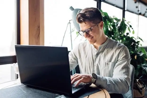 A man wearing glasses is focused on his laptop, engaged in work at a desk.