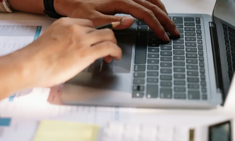 A person focused on typing on a laptop computer while seated at a desk, surrounded by a tidy workspace.