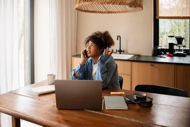 A woman seated at a table, working on a laptop while using her phone, focused and engaged in her tasks.