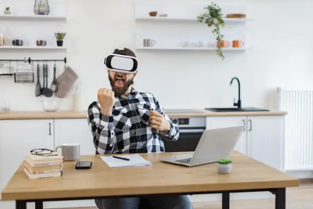 A man in a virtual reality headset sits at a table, surrounded by a laptop and a book, exploring a virtual environment.