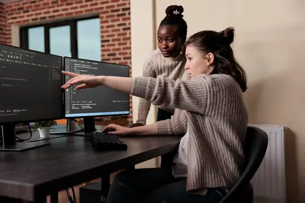 Two women collaborate on a computer, focused on their work, with a textured brick wall in the background.