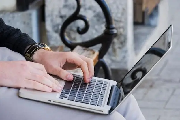 A person seated on a bench, focused on their laptop, surrounded by a serene outdoor environment.