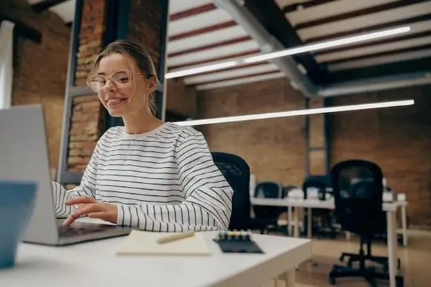 A woman wearing glasses is focused on her laptop, engaged in her work with a determined expression.