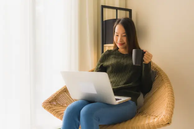 A woman seated on a chair, working on her laptop while enjoying a cup of coffee beside her.