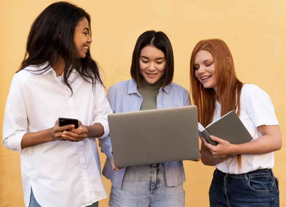 Three young women stand together, engaged with a laptop, sharing ideas and collaborating in a friendly atmosphere.